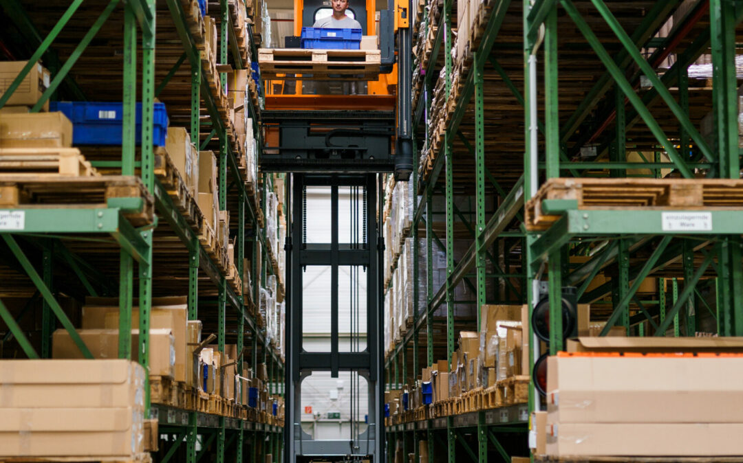 Eine Person fährt mit einem Halemeier-Kommissioniergabelstapler zwischen hohen Regalen mit gestapelten Kartons in einem Lagergang.