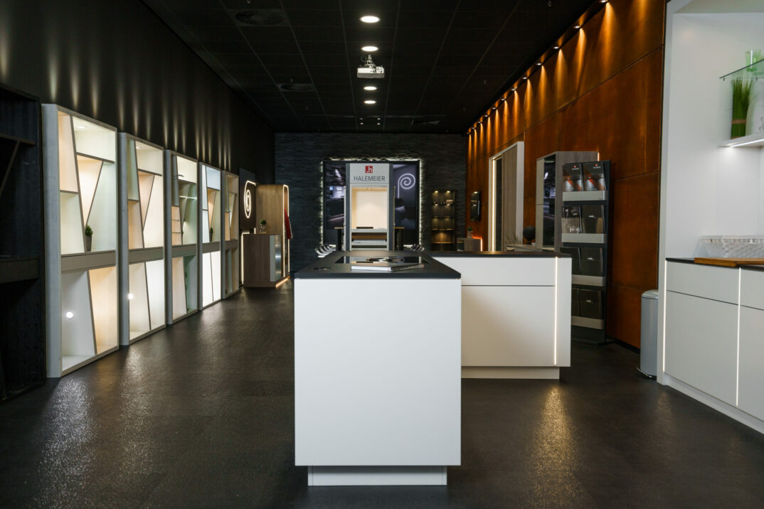 Ein moderner Ausstellungsraum mit schlichten, minimalistischen Küchenauslagen, weißen Inseln, beleuchteten Halemeier-Regalen und dunklen Böden unter Deckenstrahlern.