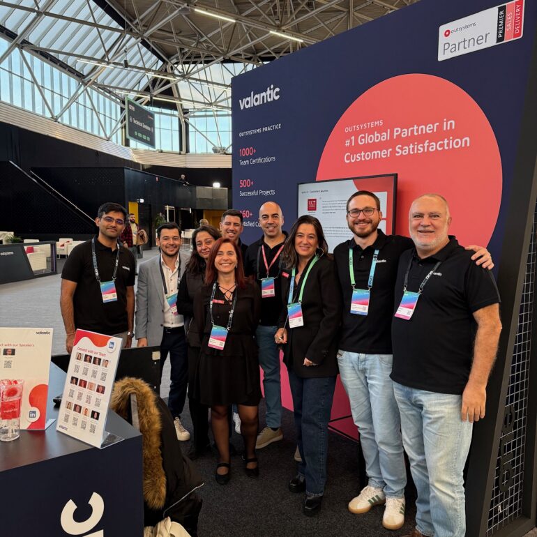 A group of eight people pose and smile at a trade show booth for Valantic, an OutSystems partner, during the ONE Conference in a convention center setting.