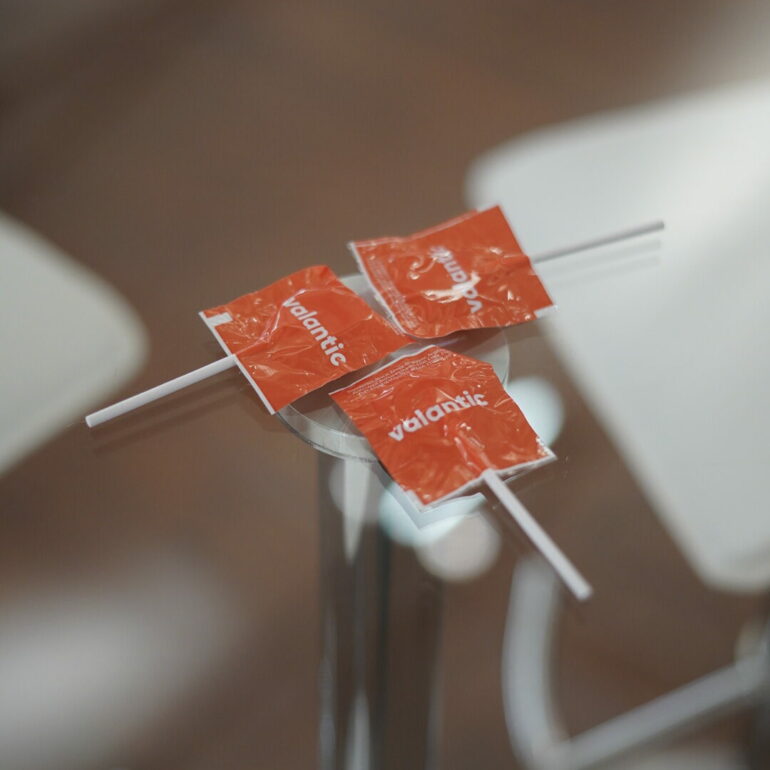 Three red lollipops in wrappers are placed on a glass table, with white chairs in the background—perfect treats for attendees gathering at the OutSystems Global ONE Conference.