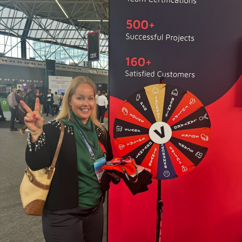A smiling woman stands beside a prize wheel and a banner showcasing OutSystems Practice achievements at the OutSystems Global ONE Conference.