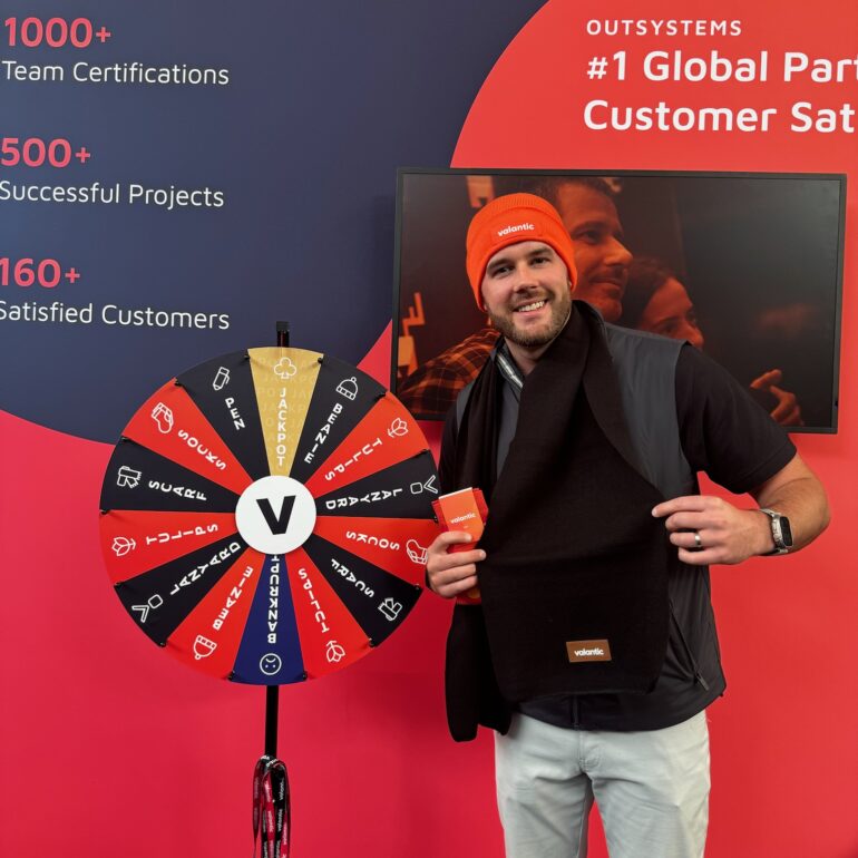 A man stands next to a prize wheel, holding a black scarf and a small orange box, wearing an orange beanie, in front of a branded ONE Lisbon promotional display.