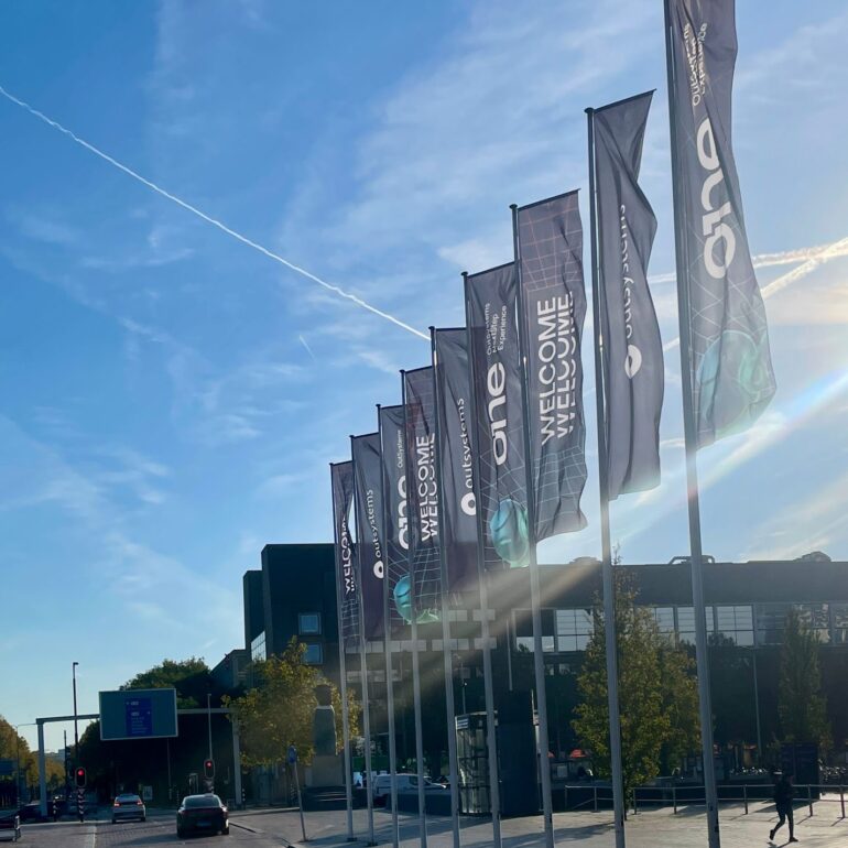A row of tall flagpoles with "One Welcome" flags line a street near a modern building under a blue sky, celebrating the OutSystems Global ONE Conference.