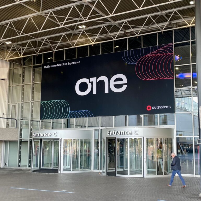A large digital sign reading "one" and "OutSystems NextStep Experience" towers above the revolving doors at Entrance C of a modern building during the OutSystems Global ONE Conference, as a person walks by.