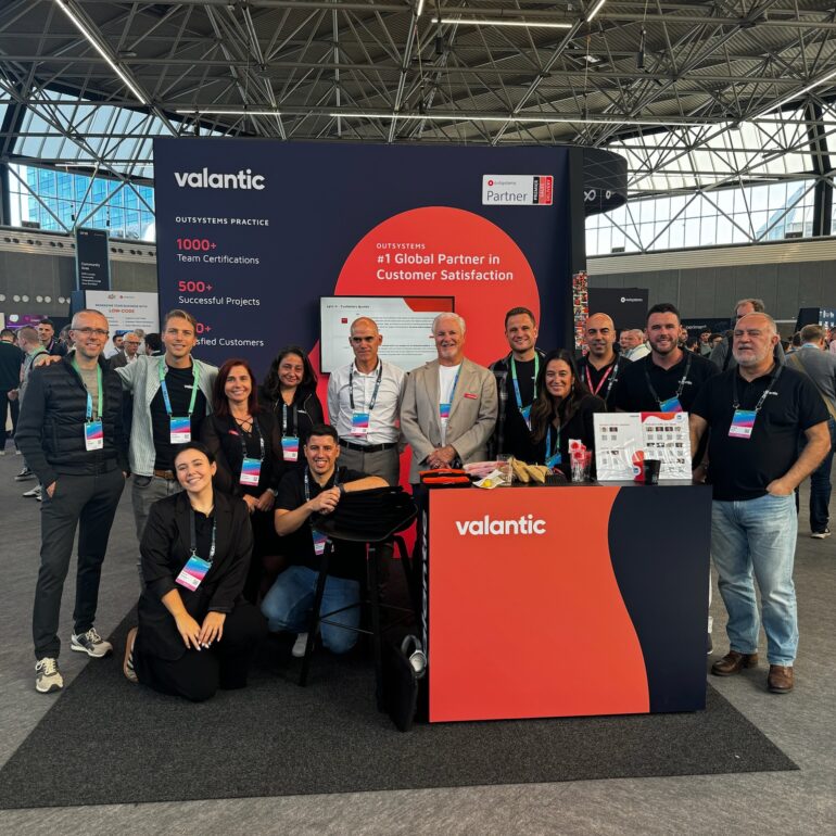 A group of people pose and smile in front of a Valantic booth at the OutSystems Global ONE Conference, with a display highlighting their customer satisfaction achievements.