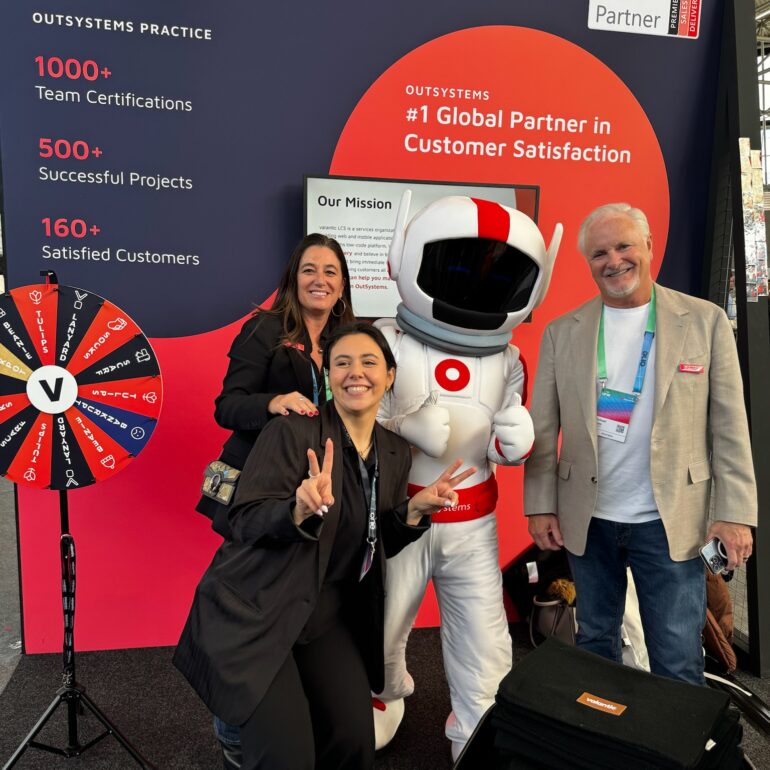 Three people pose and smile next to a person in an astronaut costume at the valantic trade show booth during the OutSystems Global ONE Conference, featuring promotional banners and a prize wheel.