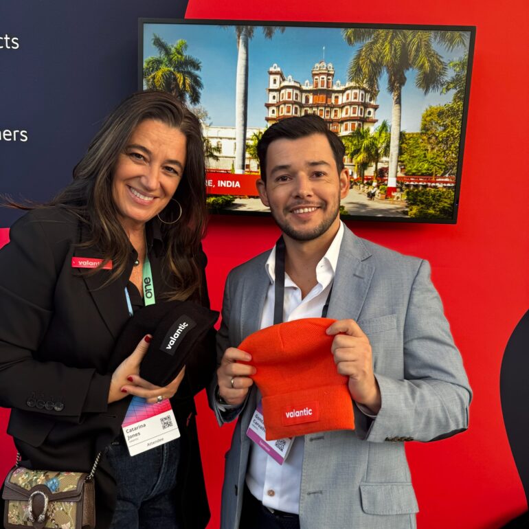 Two people stand in front of a red and black event backdrop, smiling and holding branded Volantic beanies, one black and one orange, at the OutSystems Global ONE Conference.