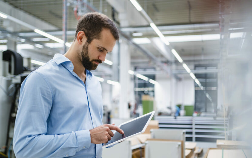 Man in a light blue shirt using a tablet in a modern industrial workspace with shelves and equipment in the background.