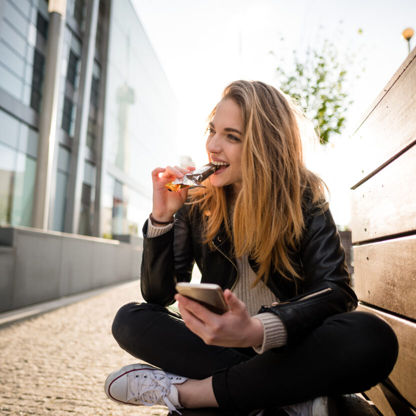 Woman eating chocolate on phone outdoor in street sitting on bench