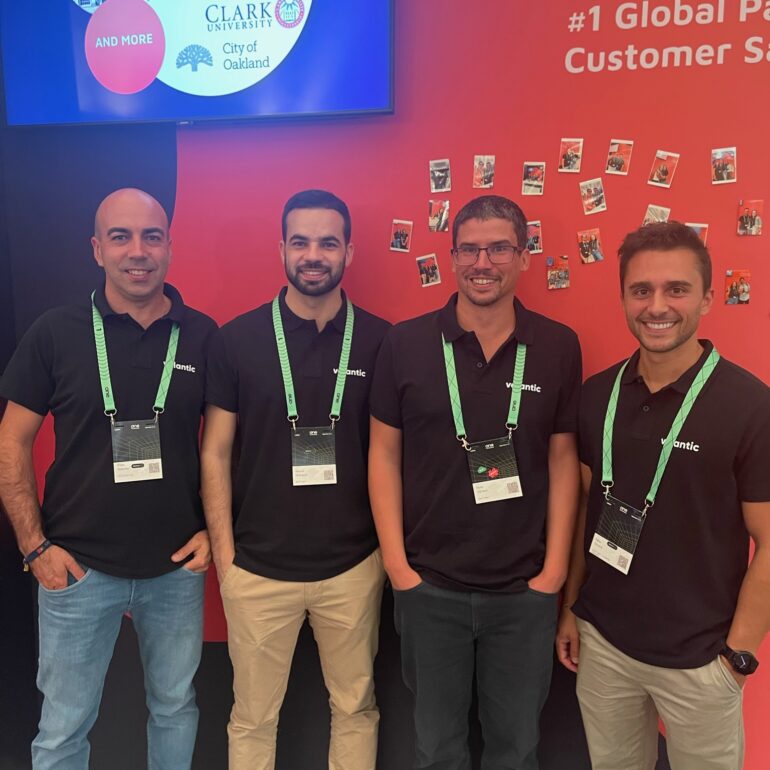 Four men wearing black polo shirts and conference badges stand in front of a red booth wall with logos and photos displayed at the OutSystems Global ONE Conference.