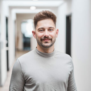 Ein Mann mit kurzen braunen Haaren und Schnurrbart steht in einem hellen Flur, trägt ein hellgraues langärmeliges Hemd und schaut in die Kamera.