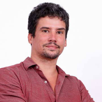 Man with short dark hair and facial hair wearing a red button-up shirt, standing against a plain white background with arms crossed, ready to attend the OutSystems Global ONE Conference.