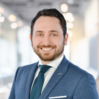 Ein Mann in blauem Anzug und Krawatte lächelt in einem modernen, hell erleuchteten Büro in die Kamera, bereit, sein Unternehmen auf dem DSAG-Jahreskongress 2025 zu vertreten.