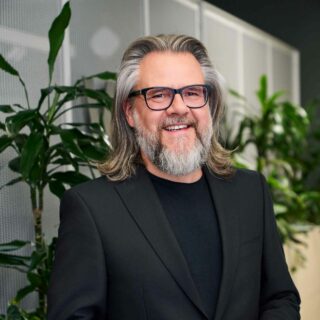 Ein Mann mit grauen Haaren und Bart, der eine Brille und einen schwarzen Anzug trägt, steht lächelnd in einer Büroumgebung mit Grünpflanzen im Hintergrund und ist bereit, am DSAG-Jahreskongress 2025 teilzunehmen.