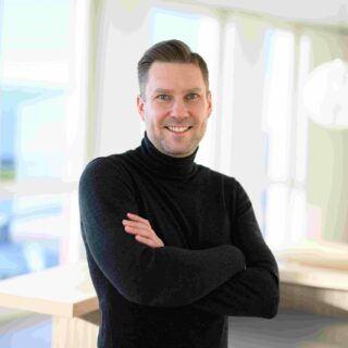 Ein Mann mit kurzen Haaren und schwarzem Rollkragenpullover steht mit verschränkten Armen in einem Innenraum und lächelt in die Kamera. Die helle, moderne Einrichtung deutet auf ein professionelles Umfeld wie den DSAG-Jahreskongress 2025 hin.