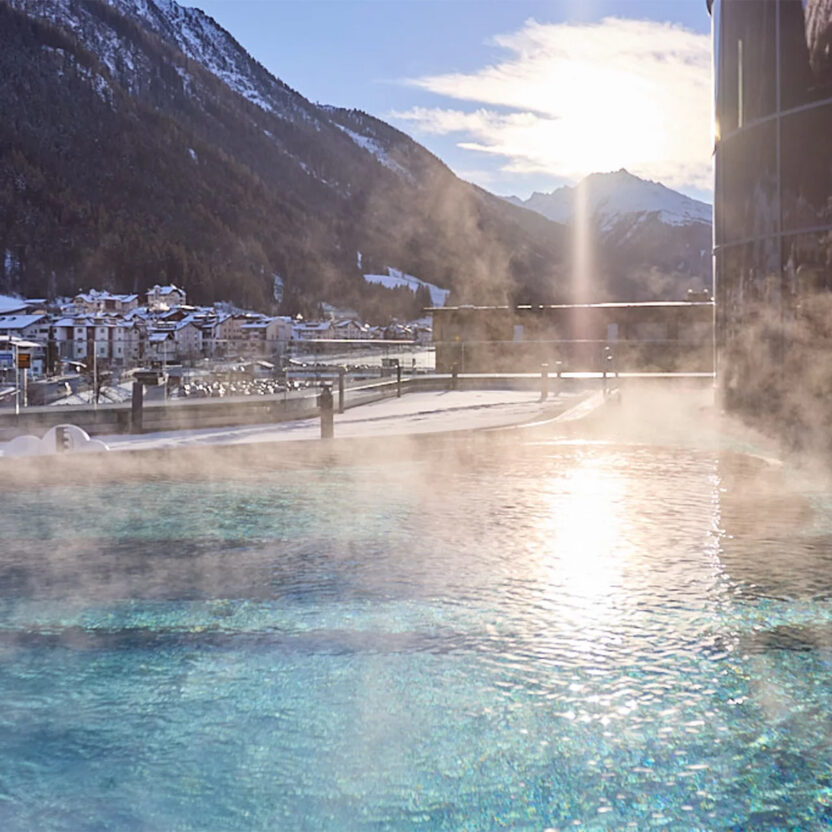Outdoor pool of the Silvretta Therme Ischgl with a view of wintry Ischgl and the surrounding mountain landscape at sunrise.