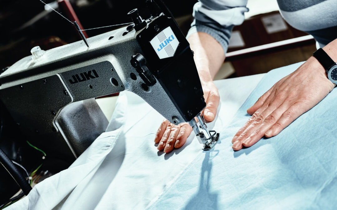 A person guides light blue fabric through an industrial Juki sewing machine, stitching a seam while wearing a watch.
