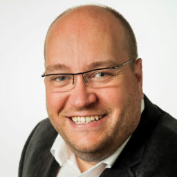 Smiling man with glasses and a receding hairline, wearing a dark suit jacket and white shirt, photographed against a plain white background.