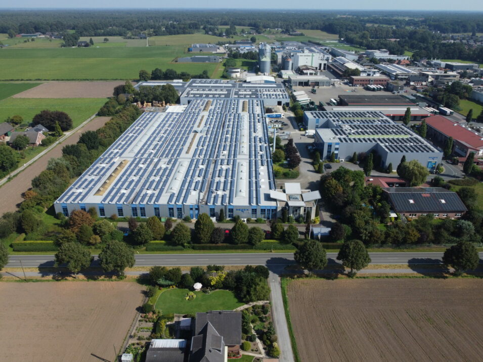 Aerial view of a large industrial complex surrounded by fields and trees, with multiple buildings and parking areas visible.
