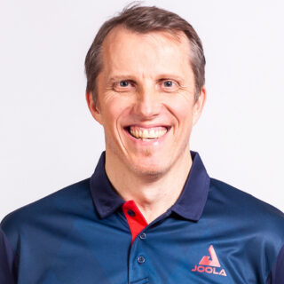 A smiling man with short brown hair wears a navy blue collared shirt with a red accent and the JOOLA logo on the chest at DEF26, the Digital Excellence Forum.