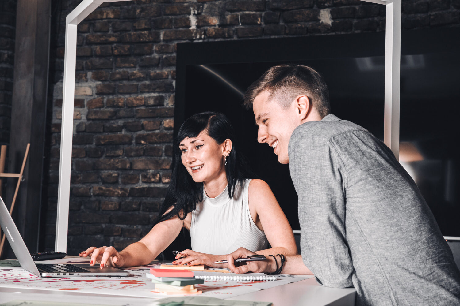 Two people are sitting at a desk in an office, looking at a laptop screen and smiling.