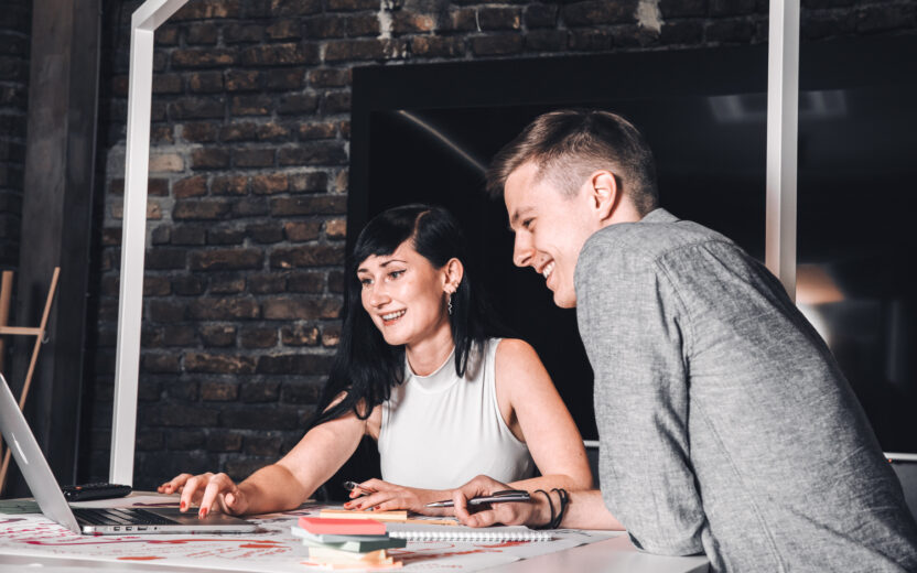 Two people are sitting at a desk in an office, looking at a laptop screen and smiling.