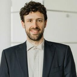 Ein Mann mit lockigem dunklem Haar und Bart, der eine dunkle Anzugsjacke und ein helles Hemd trägt, steht in einem Haus und lächelt in die Kamera.