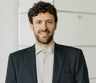 Ein Mann mit lockigem dunklem Haar und Bart, der eine dunkle Anzugsjacke und ein helles Hemd trägt, steht in einem Haus und lächelt in die Kamera.