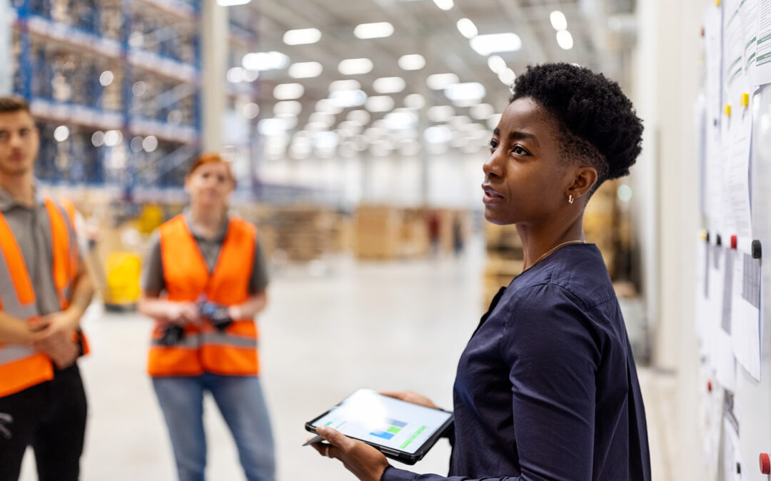 Supervisor standing by a whiteboard with a digital tablet discussing dispatch plan with workers. Team of workers having meeting in a distribution warehouse.