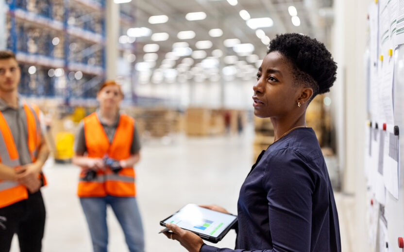 Supervisor standing by a whiteboard with a digital tablet discussing dispatch plan with workers. Team of workers having meeting in a distribution warehouse.