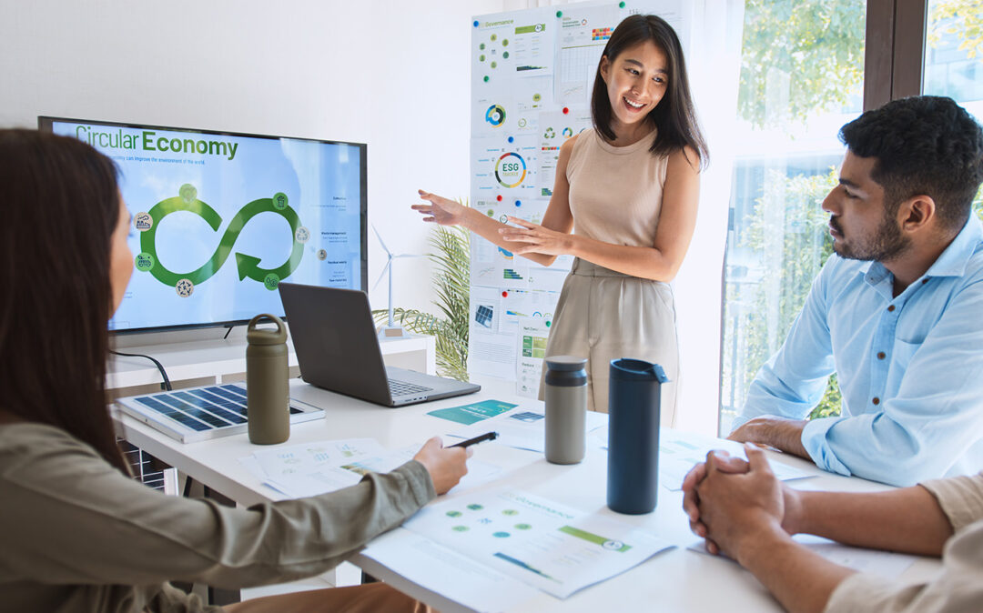 Geschäftsfrau leitet Teambesprechung und zeigt auf einem Monitor Präsentation zu Nachhaltigkeit.