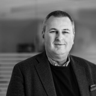 Ein Mann mittleren Alters in Anzugjacke und Pullover steht in einem Haus und blickt mit einem leichten Lächeln in die Kamera, als würde er darüber nachdenken, was wäre wenn" - der Hintergrund ist mit horizontalen Linien verschwommen.