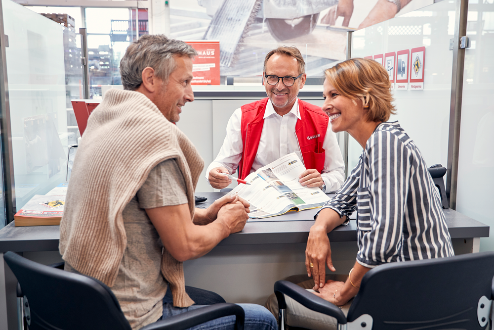 Herr berät ein Paar über Montagen. Alle drei sitzen an einem Tisch mit einem offenen Magazin. Das Paar ist im Gespräch.