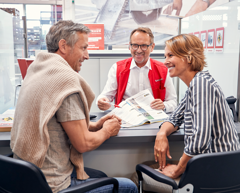 Herr berät ein Paar über Montagen. Alle drei sitzen an einem Tisch mit einem offenen Magazin. Das Paar ist im Gespräch.
