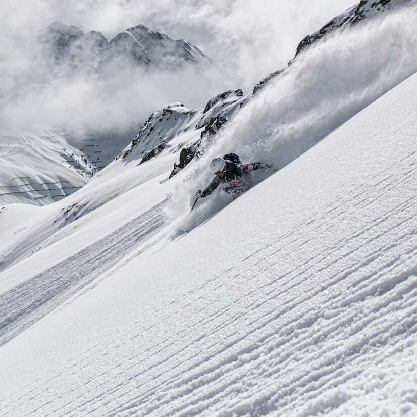 Ein Skifahrer fährt einen steilen, schneebedeckten Hang der Silvretta Montafon hinunter und wirbelt dabei Pulverschnee auf, während im Hintergrund schroffe Gipfel und Wolken zu sehen sind.