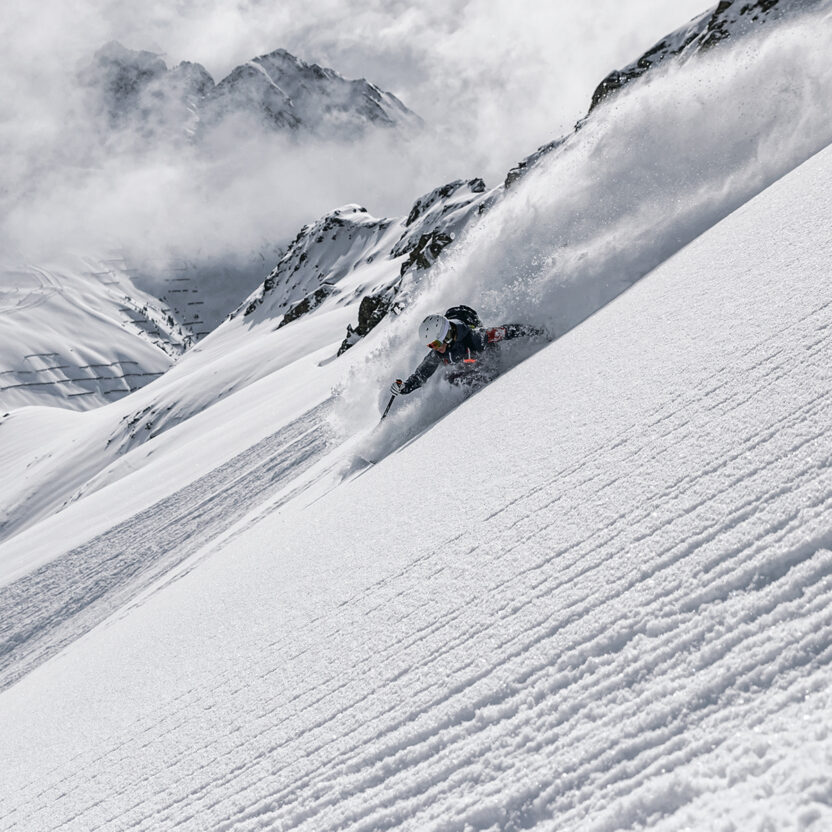 Ein Skifahrer fährt einen steilen, schneebedeckten Hang der Silvretta Montafon hinunter und wirbelt dabei Pulverschnee auf, während im Hintergrund schroffe Gipfel und Wolken zu sehen sind.