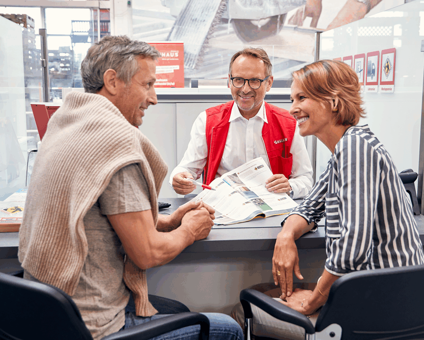Herr berät ein Paar über Montagen. Alle drei sitzen an einem Tisch mit einem offenen Magazin. Das Paar ist im Gespräch.