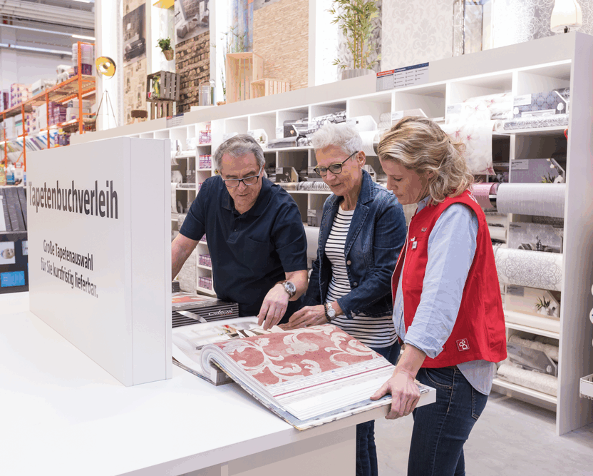 Dame berät ein Ehepaar bei Tapetenmustern. Buch mit Vorlagen auf dem Tisch und davor Beschreibungsdesk mit Aufschrift Tapetenbuchverleih