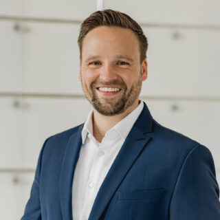 Ein Mann in dunkelblauem Anzug und weißem Hemd steht in einem Innenraum und lächelt in die Kamera vor einem unscharfen, hellen Hintergrund, der das Vertrauen in die ESG-Umsetzung und eine effektive ESG-Roadmap widerspiegelt.