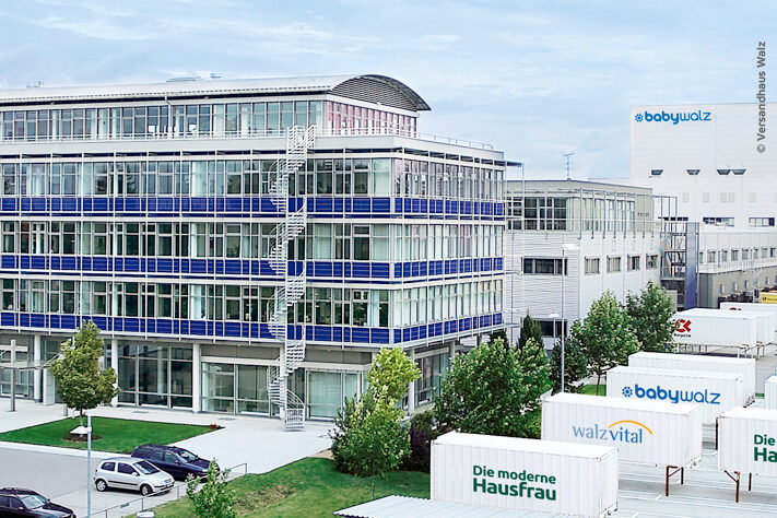 Moderne Büro- und Lagergebäude mit "babywalz"-Schildern, parkende Autos, Bäume und Schiffscontainer mit der Aufschrift "walz vital" und "Die moderne HausFrau" spiegeln einen Standort wider, der die ESG-Umsetzung in seinen täglichen Betrieb integriert.