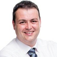 Mann mit kurzen dunklen Haaren, weißem Hemd und gestreifter Krawatte, der in die Kamera lächelt, vor einem schlichten weißen Hintergrund - bereit, Sie bei der ESG-Umsetzung oder Nachhaltigkeitsberichterstattung zu unterstützen.