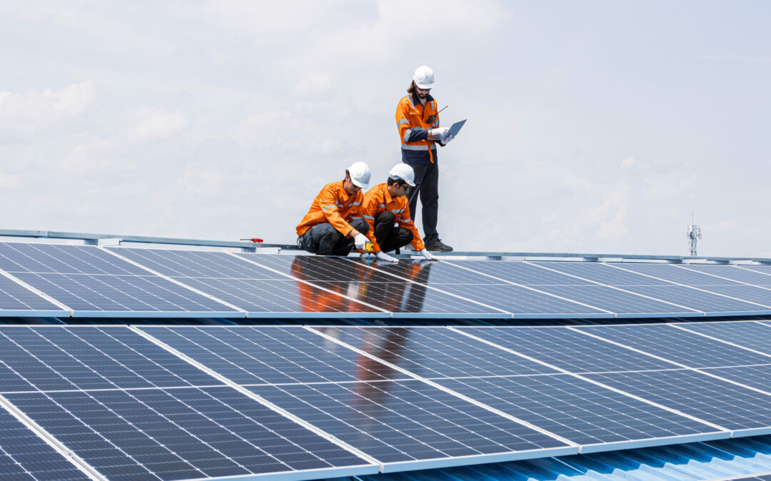 Drei Arbeiter in orangefarbenen Sicherheitsuniformen und Helmen installieren und überprüfen unter freiem Himmel Solarpaneele auf einem Dach und unterstützen Energieversorger mit intelligenten Lösungen für die Digitalisierung.