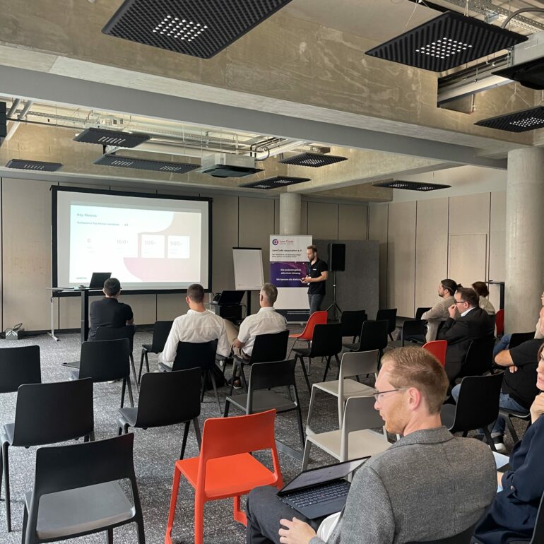 A presenter stands at the front of a conference room giving a presentation to attendees seated in rows, with a slide projected on a screen at the 2025 German Low-Code Day.