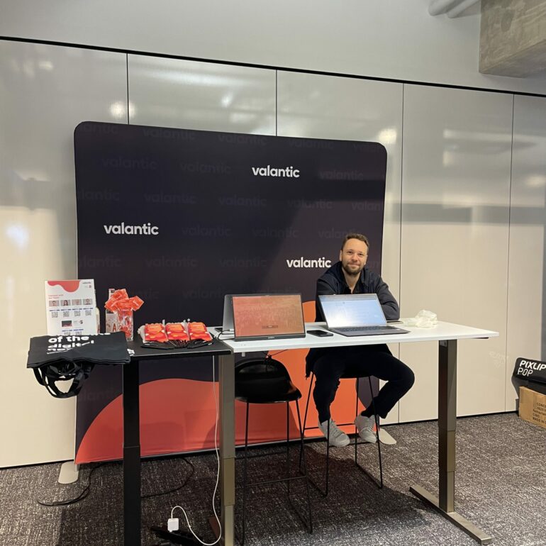 A man sits behind a display table with laptops and promotional items in front of a valantic-branded backdrop at the 2025 German Low-Code Day indoor event.
