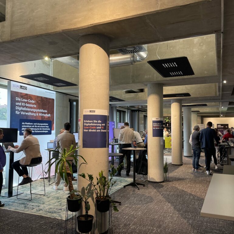 Attendees gather at the 2025 German Low-Code Day in a modern indoor space, surrounded by concrete pillars, informational banners, and potted plants.