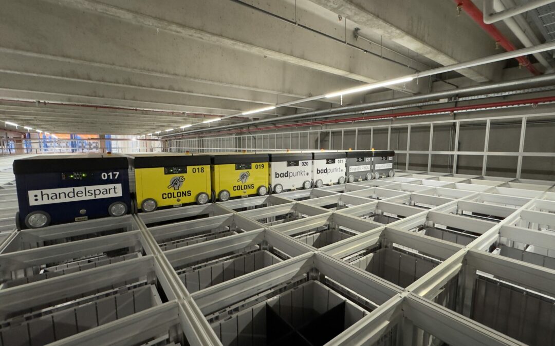 Ein automatisiertes Lager mit Robotern auf Schienen, markiert mit verschiedenen Marken. Die Roboter bewegen sich über einer gitterartigen Struktur aus offenen Fächern zur Lagerung, in einem gut beleuchteten Raum mit freiliegenden Rohren an der Decke.