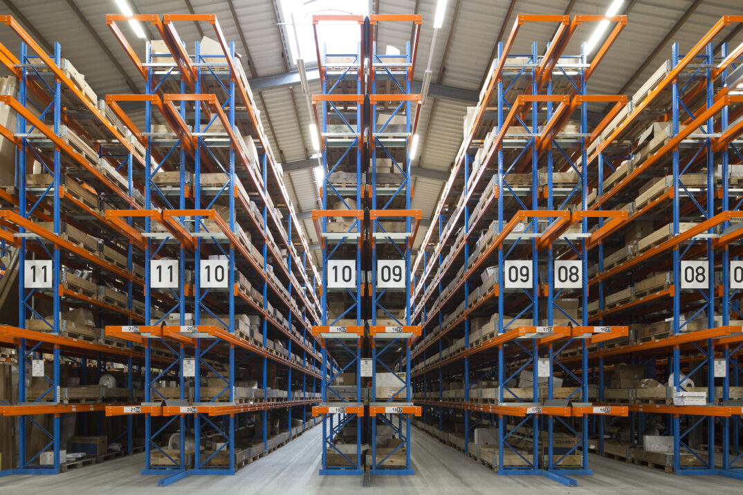 Ein großes Lager mit hohen Regalen in Blau und Orange, beschriftet mit Abschnittsnummern, gefüllt mit Kartons und Paletten. Der Raum ist hell beleuchtet und besitzt ein hohes Metalldach.