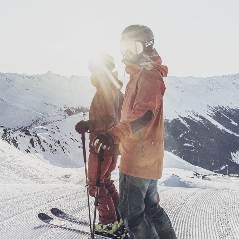 Zwei Skifahrer in Winterausrüstung stehen auf einer präparierten Davoser Skipiste, im Hintergrund schneebedeckte Berge, hinter ihnen scheint das Sonnenlicht.