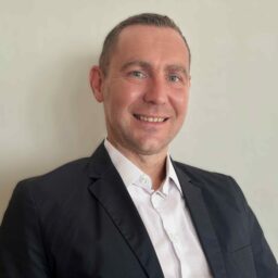 Porträt eines lächelnden Mannes in weißem Hemd und dunklem Sakko vor neutralem Hintergrund.