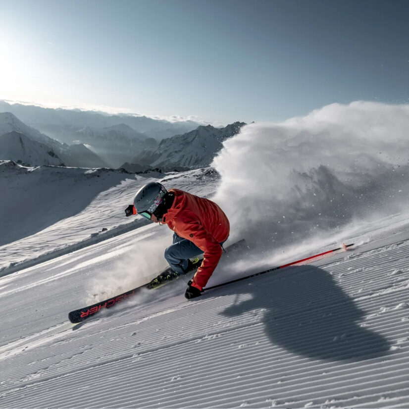 Skier in a red jacket carving down a freshly groomed slope, snow spraying up.